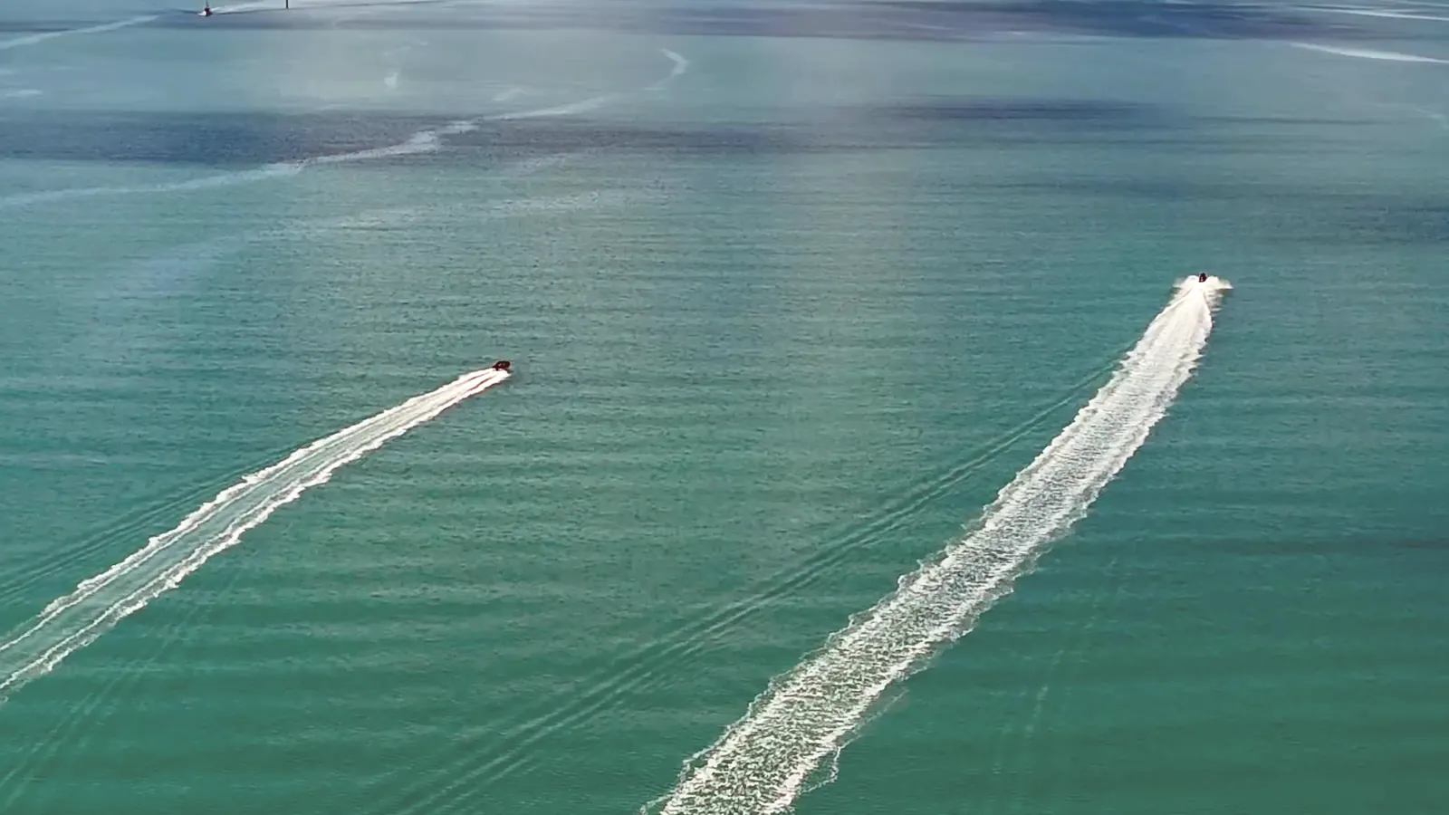 Vue du ciel : deux bateaux traversent une baie émeraude en tirant chacun un long sillage blanc.